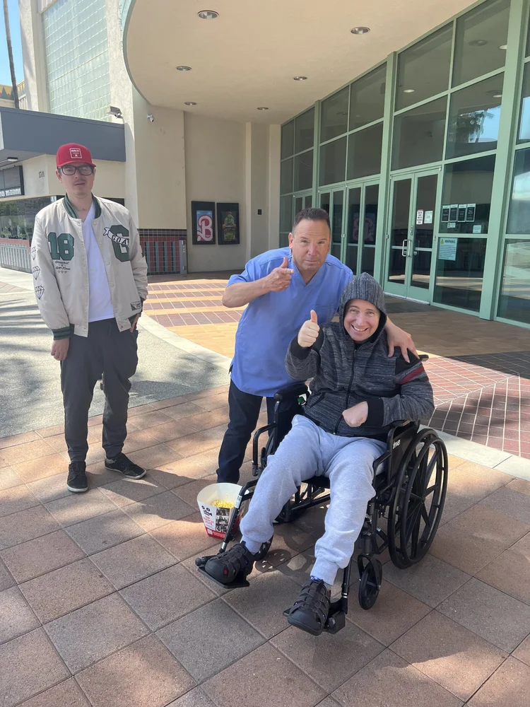 Smiling resident in wheelchair with two caregivers at facility entrance